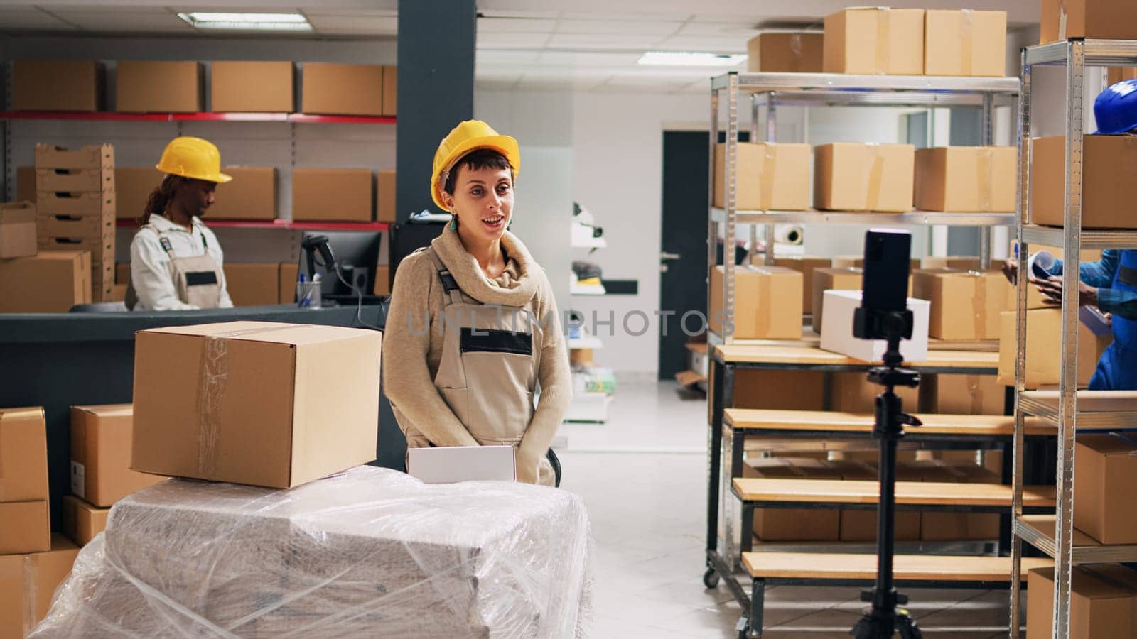 Young adult filming advertisement video in stockroom, showing products boxes on online social media page. Storage room workers using mobile phone to record PR ad, industrial goods.