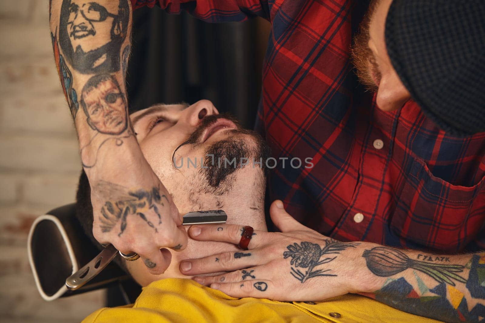 Client with beard and moustache sit on chair, and professional barber make beard shaving in barber shop