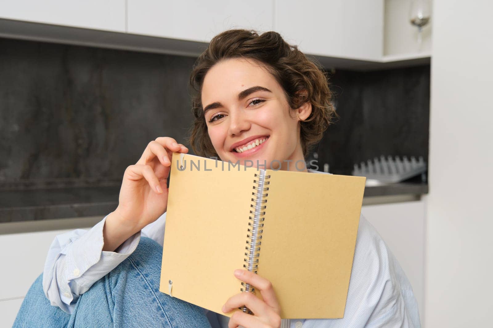 Image of brunette girl at home, holding notebook, reading through her notes, preparing for exam, doing homework in kitchen by Benzoix