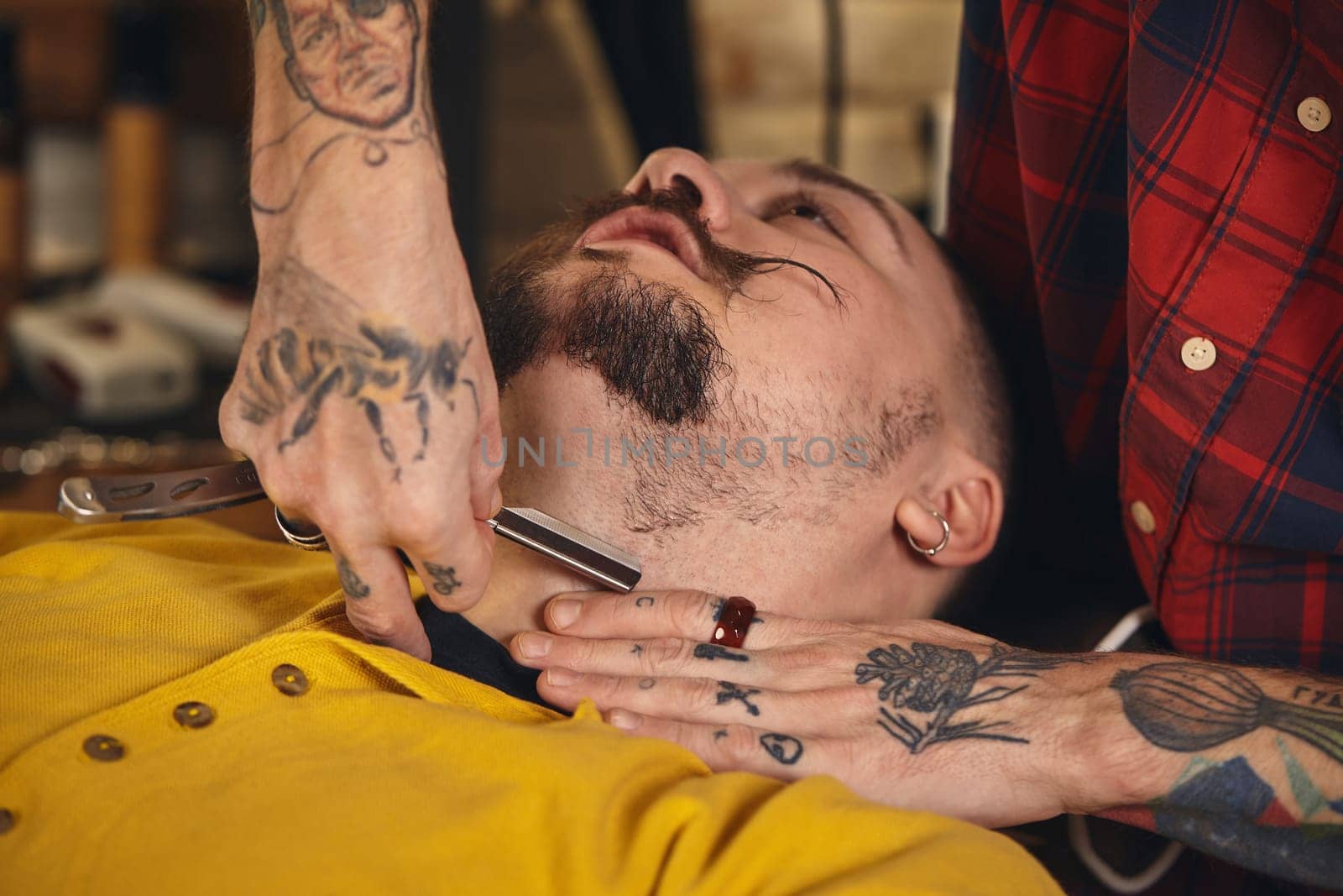 Client with beard and moustache sit on chair, and professional barber make beard shaving in barber shop