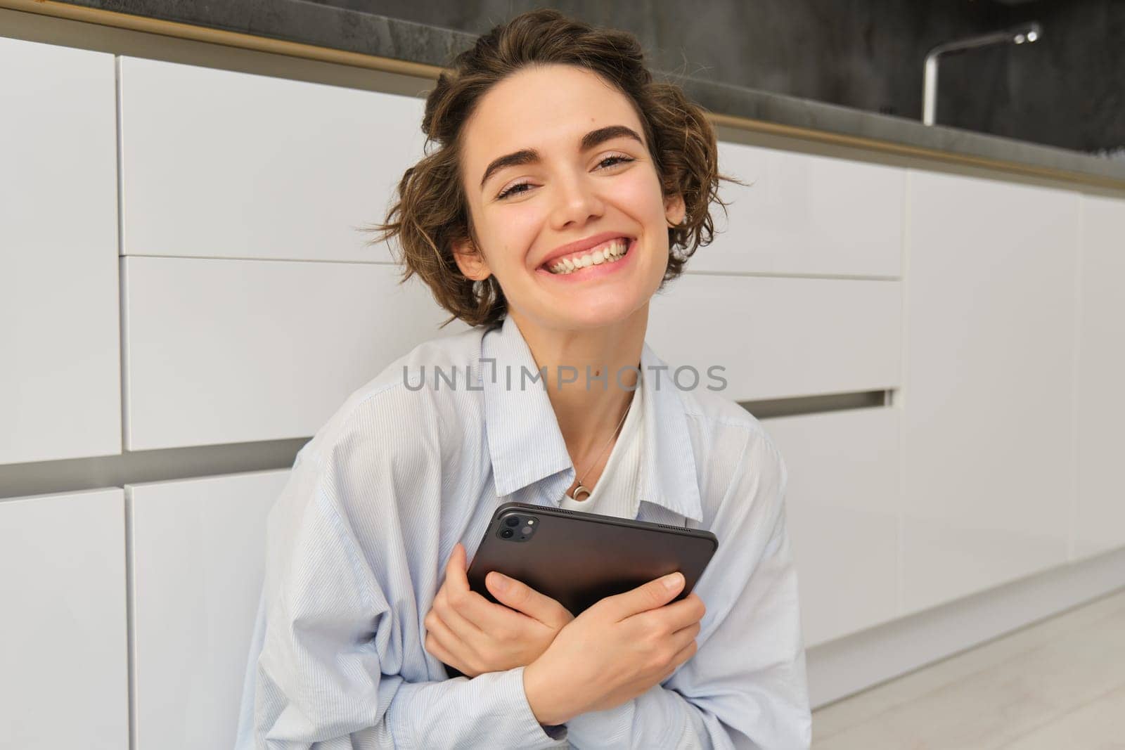 Portrait of young adult woman works on her tablet, sits with notebook on kitchen floor at home, studies in comfort.