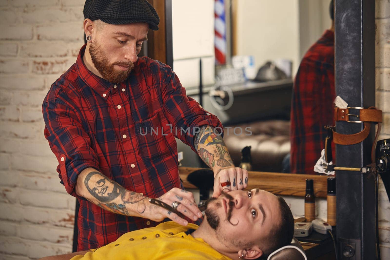 Client with beard and moustache sit on chair, and professional barber make beard shaving in barber shop