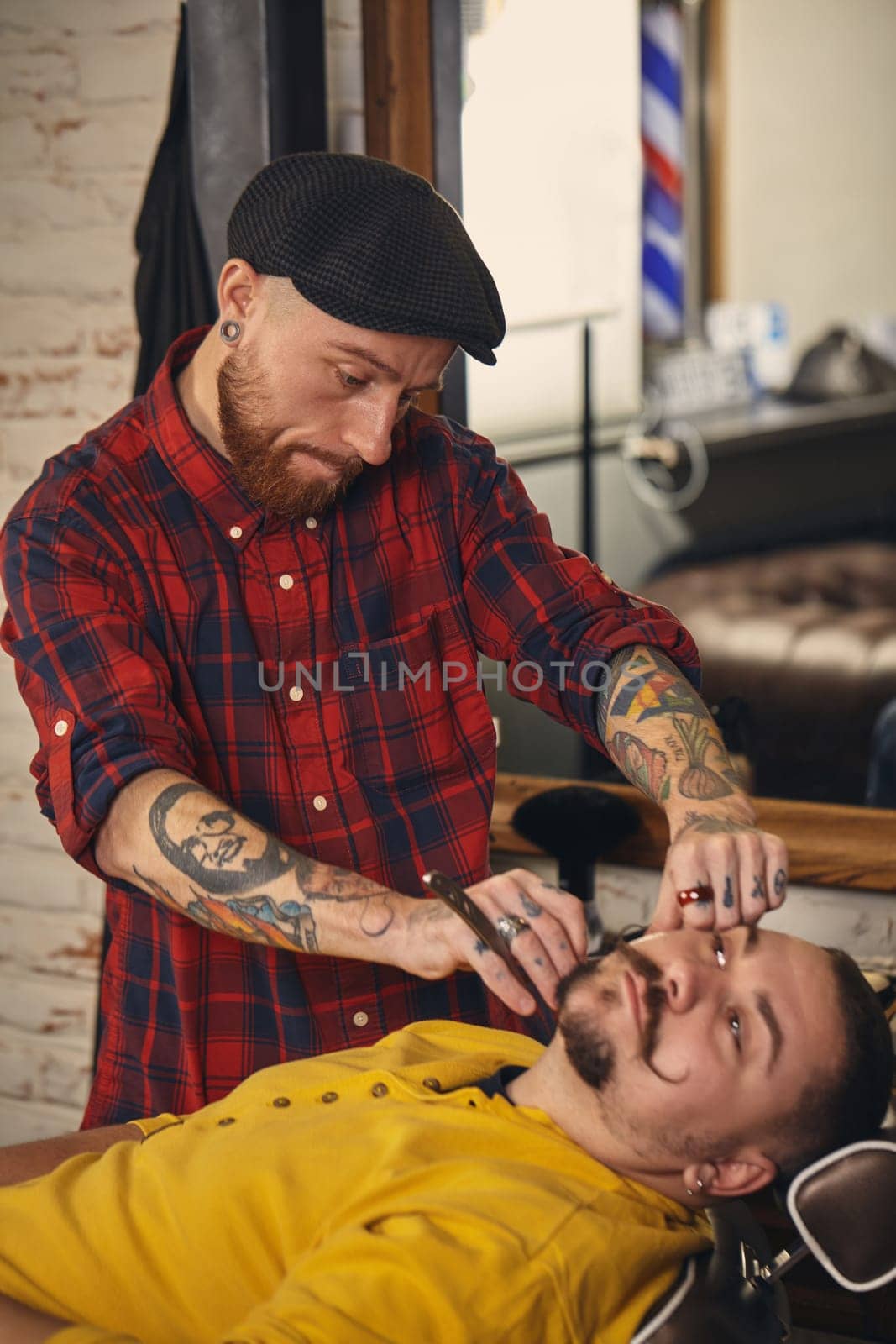 Client with beard and moustache sit on chair, and professional barber make beard shaving in barber shop