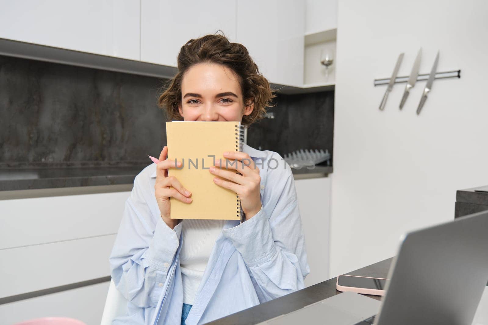 Portrait of smiling brunette girl, holds her planner, writes in notebook and looks happy, laughs, does homework, makes notes, sits at home in kitchen.