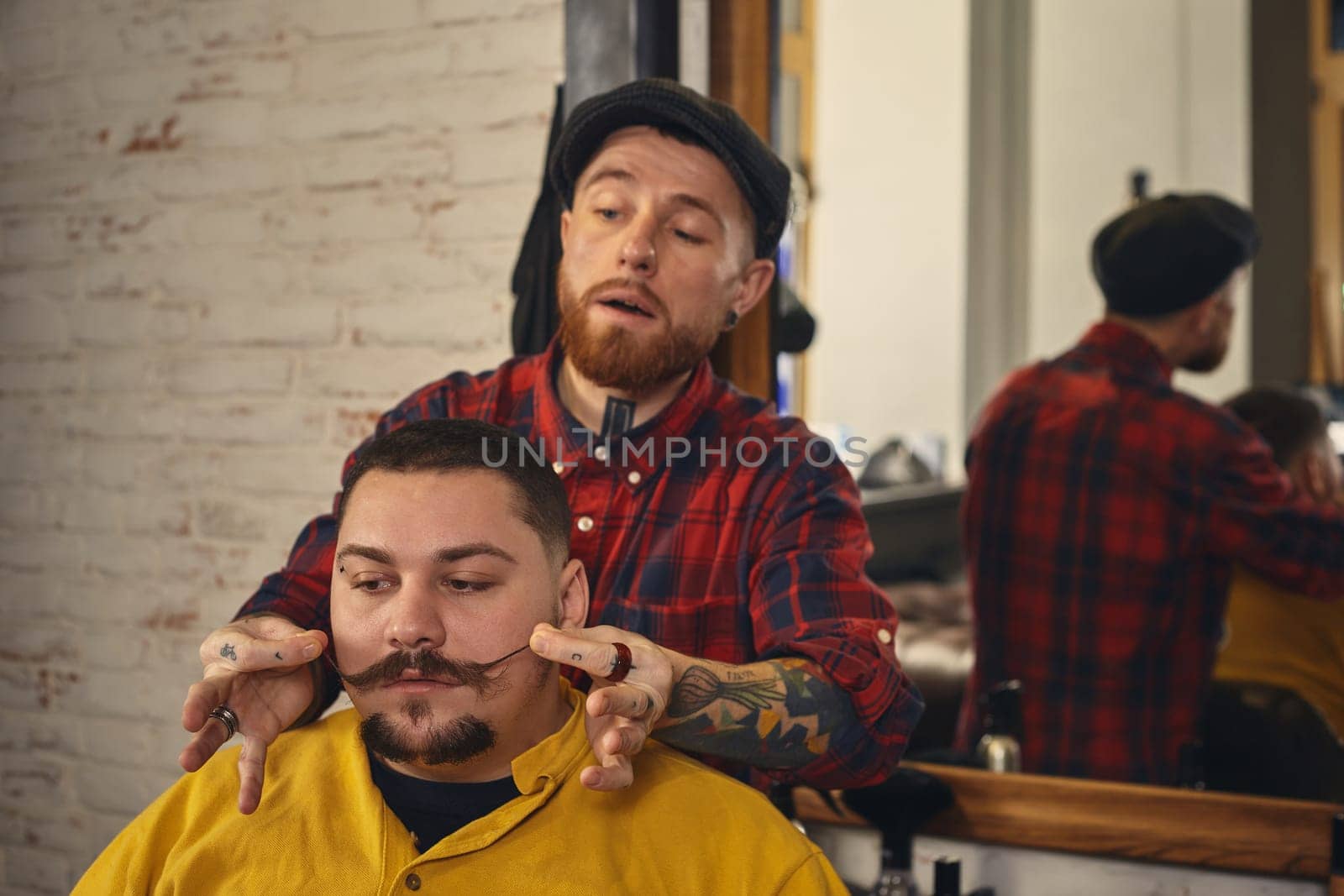 Client with beard and moustache sit on chair, and professional barber make beard shaving in barber shop. The barber straightens the mustache of the client