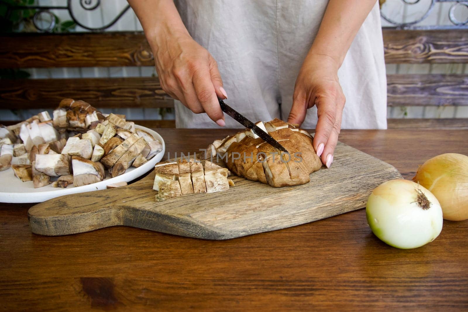 Women's hands cut wild mushrooms on a wooden board. There is a basket of mushrooms on the table and there are onions.