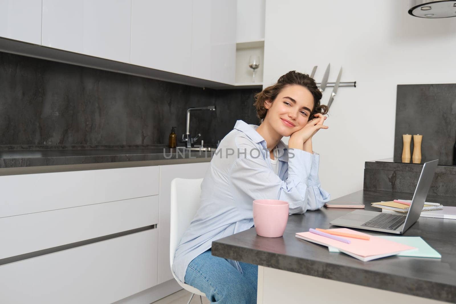 Beautiful brunette woman, looking outside window, working from home, drinking coffee in kitchen and suign laptop.