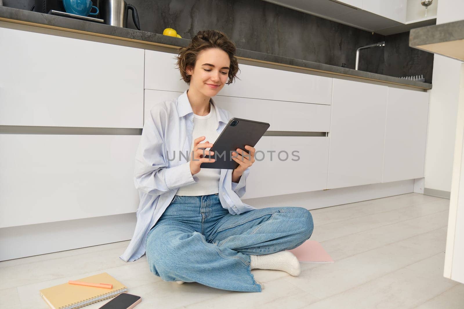 Portrait of young adult woman works on her tablet, sits with notebook on kitchen floor at home, studies in comfort by Benzoix