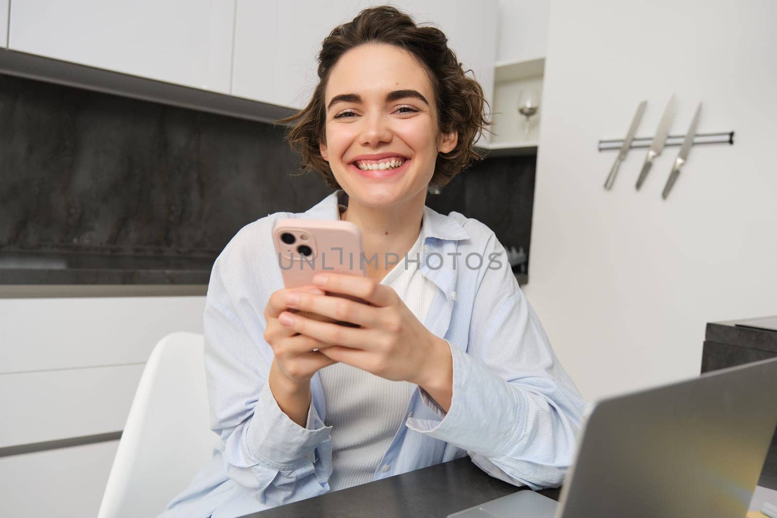 Image of young modern woman, holding mobile phone, sitting in her kitchen at home, order delivery on smartphone app.