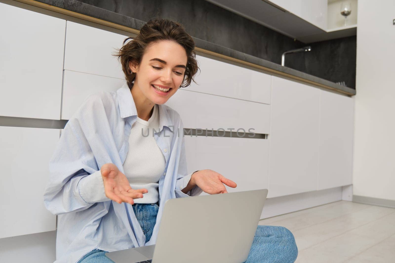 Portrait of young freelance girl connects to online meeting, video chats on laptop, sits on kitchen floor and enjoys her conversation.