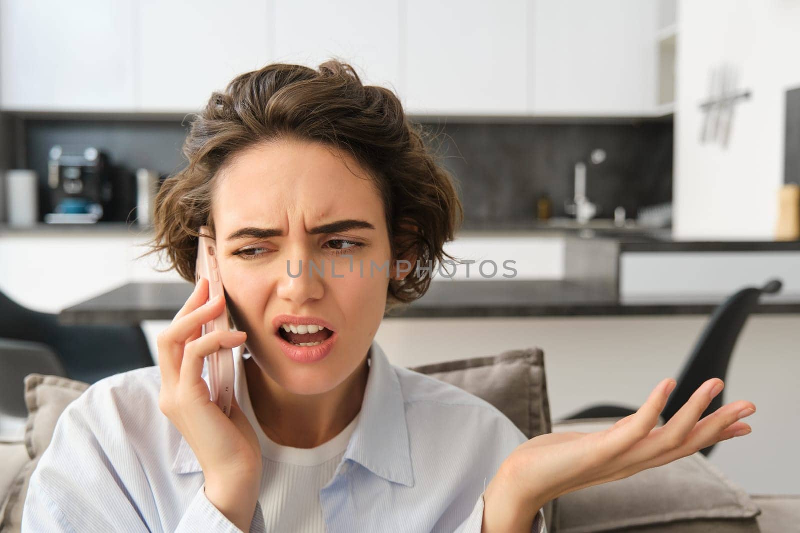 Image of woman with confused, annoyed face, talking on mobile phone while sitting at home on sofa, shrugging and looking bothered.