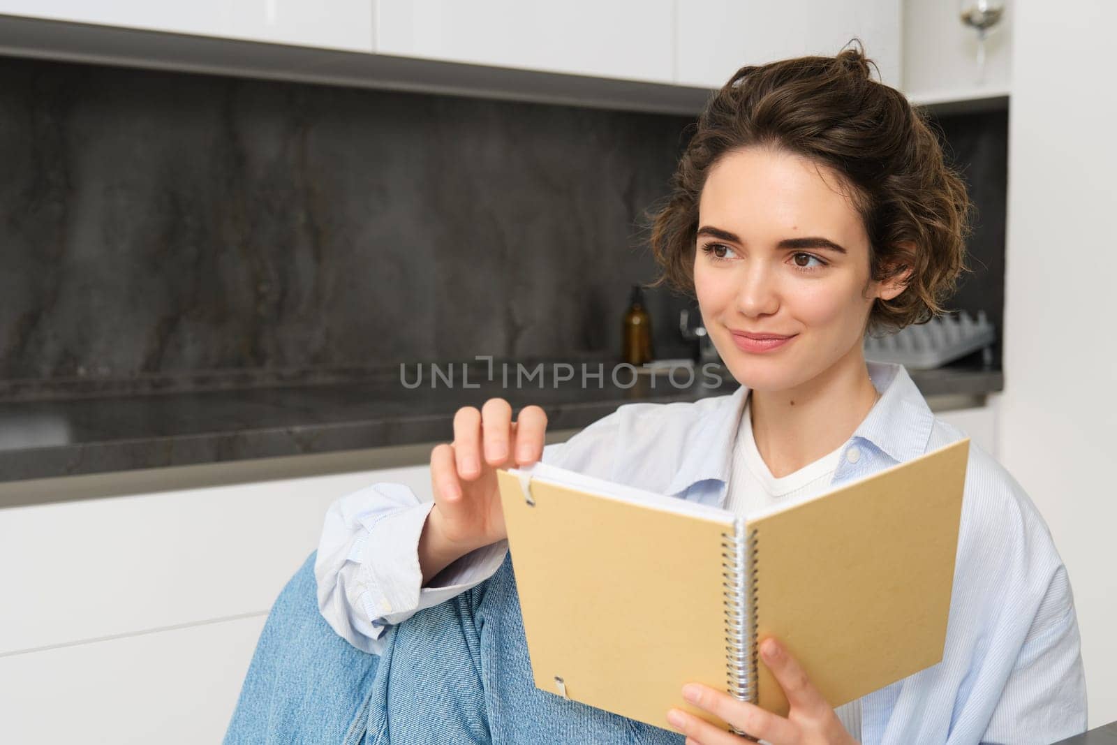 Portrait of happy smiling woman, holding notebook, studying in her kitchen, reading journal. Lifestyle and people concept