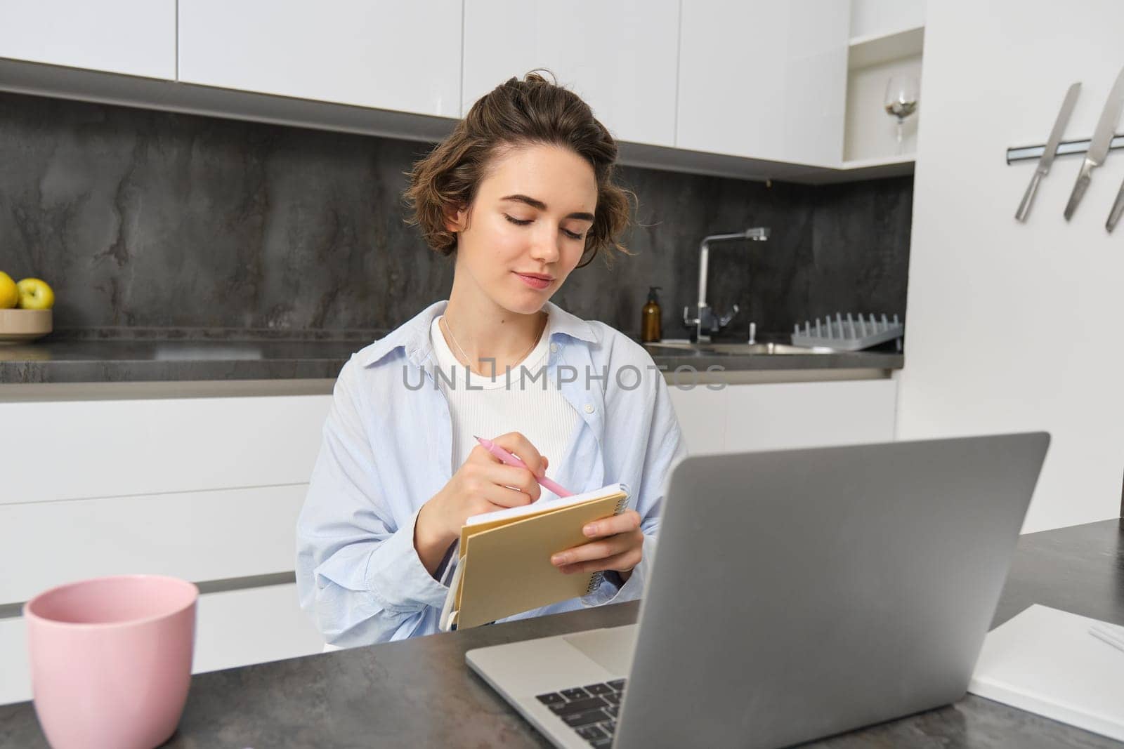 Working woman makes notes, writes down information on notebook, sitting with laptop at home.