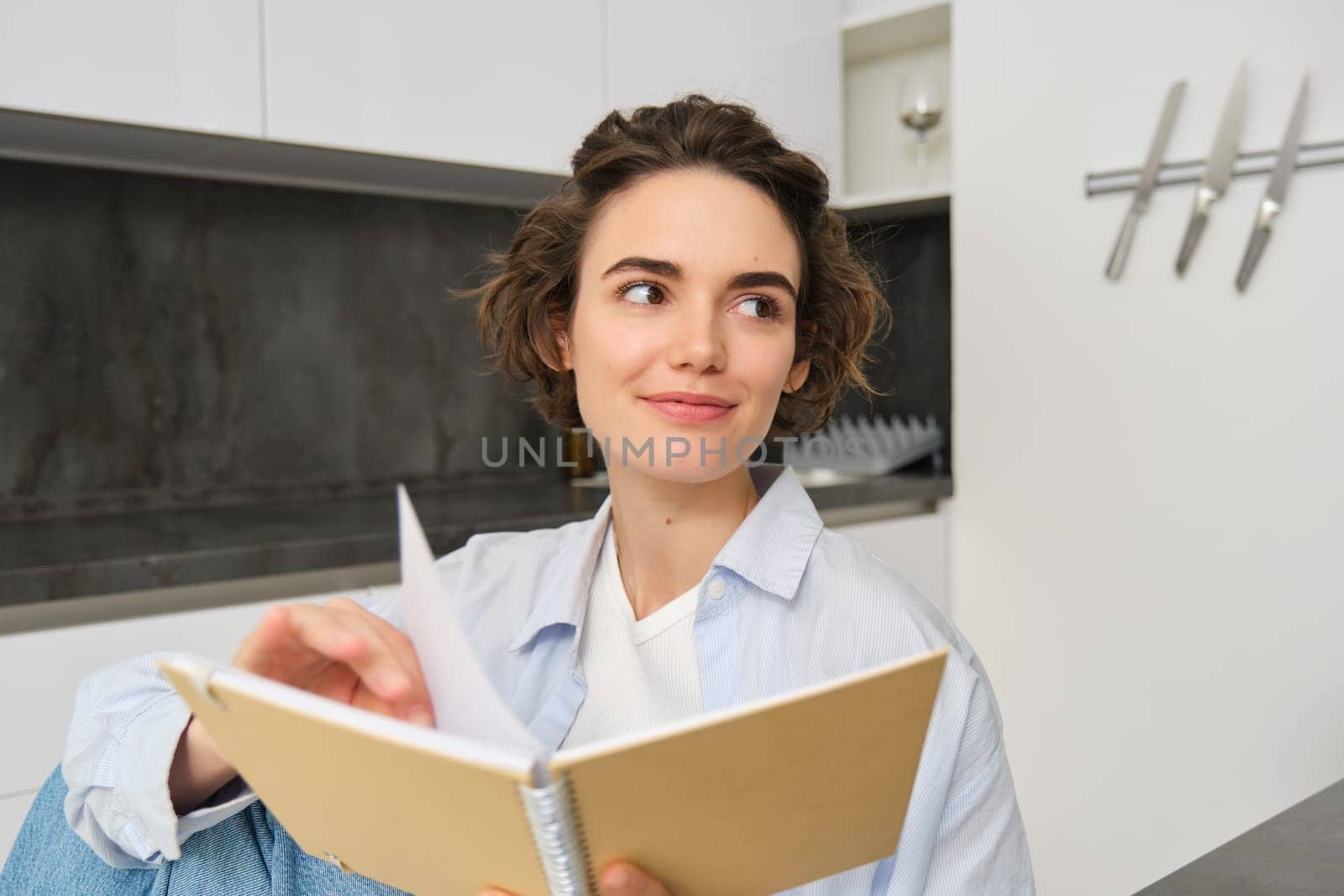 Close up portrait of modern brunette woman, holding journal, making notes in planner, reading her notebook while sitting at home in kitchen.