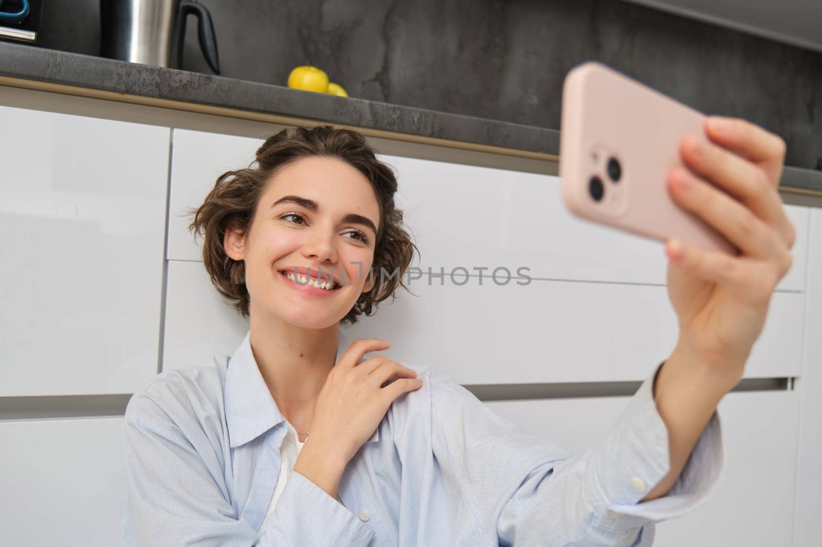 Portrait of young woman sits on kitchen floor with telephone, takes selfie on smartphone with app filters, poses for photo on mobile phone.