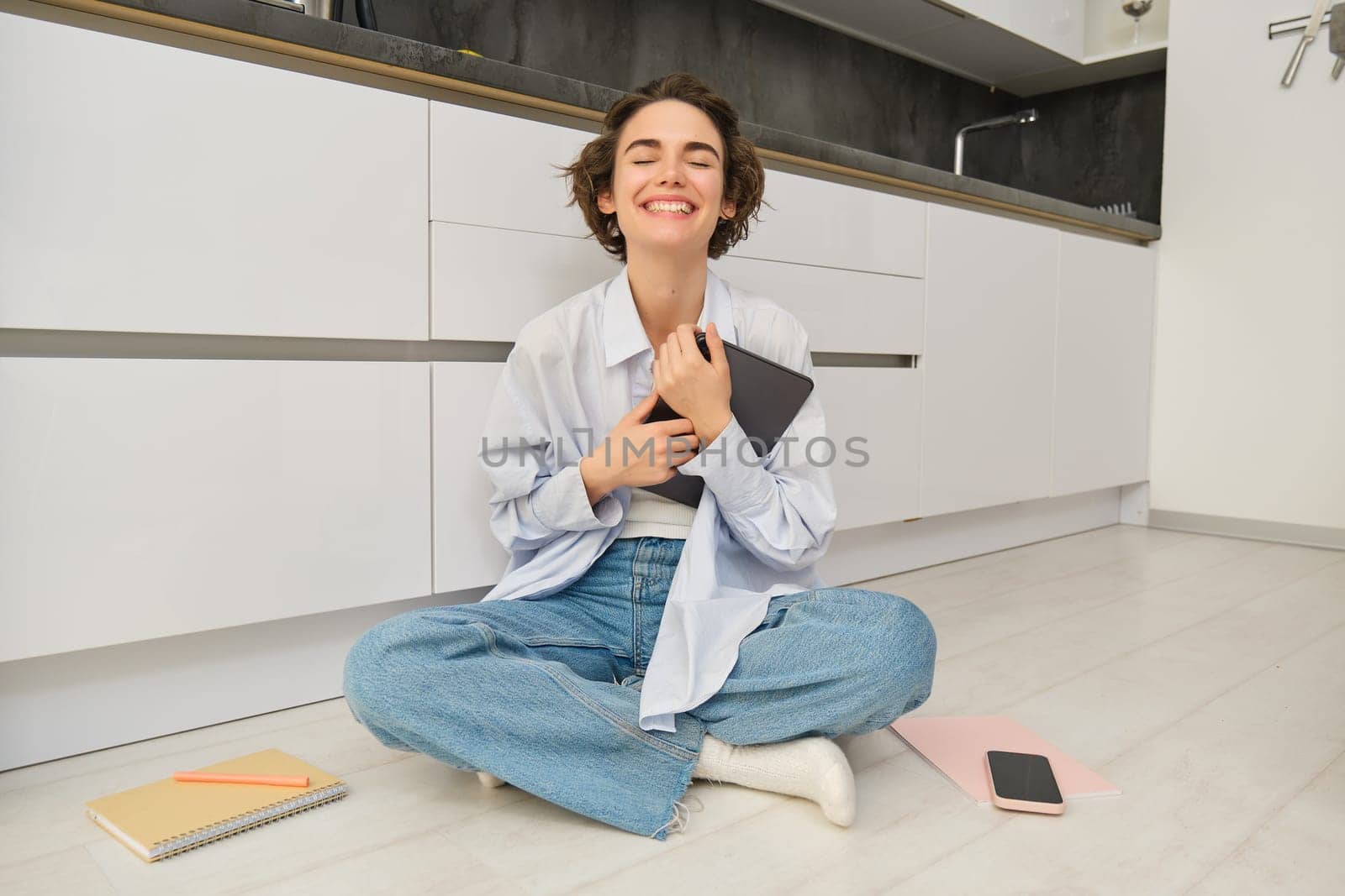 Image of happy young woman sits at home on floor, holds digital tablet, does online shopping, reading e-book. Copy space