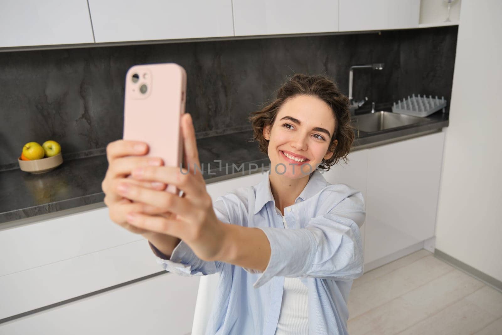 Stylish smiling girl takes selfie on smartphone at home, makes photo of herself in kitchen, poses for picture with mobile phone.