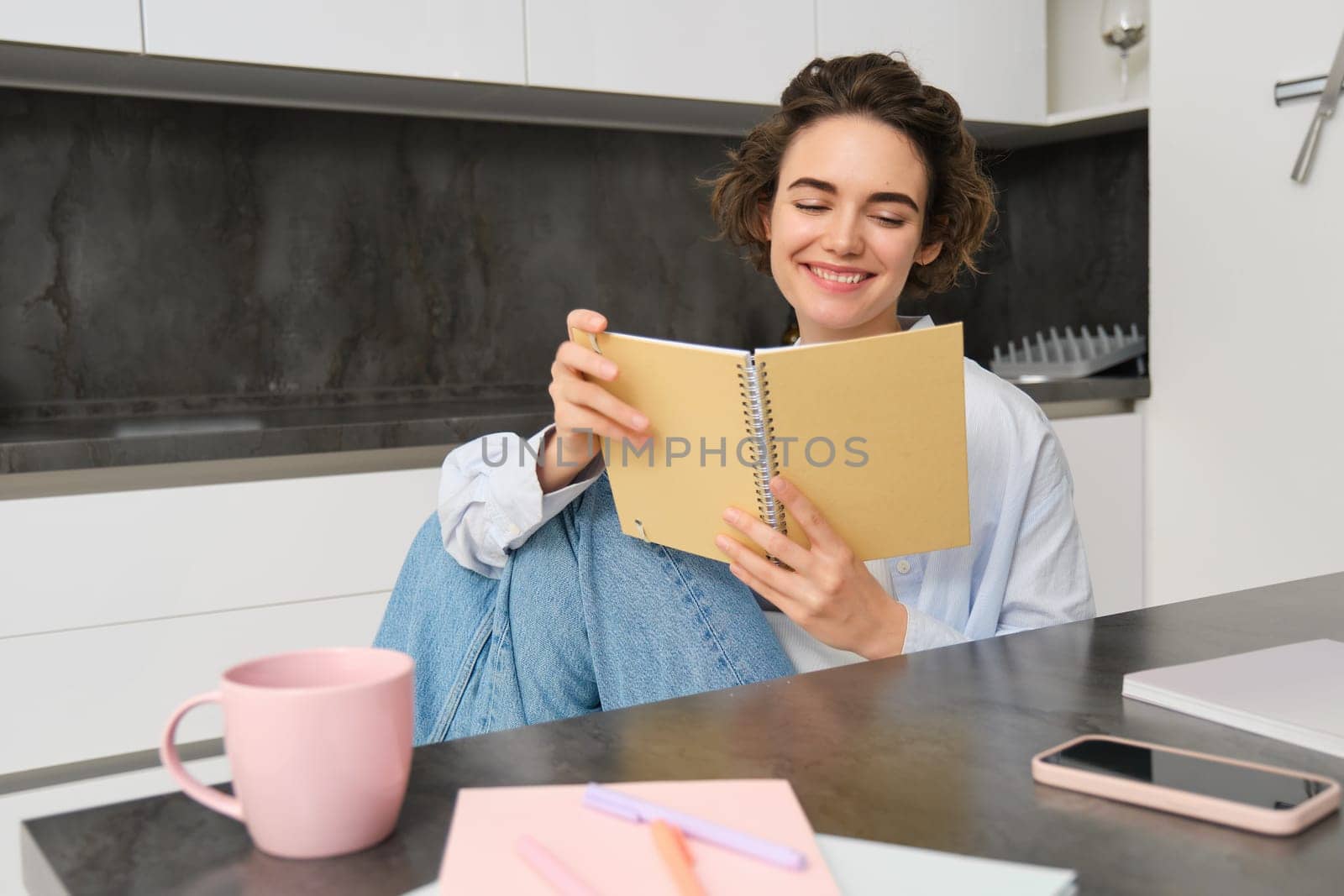 Smiling beautiful woman, sitting with notebook in kitchen, reading notes, studying, doing homework.