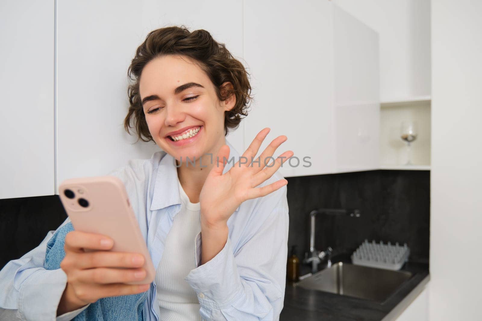 Portrait of friendly smiling brunette woman, sitting at home in kitchen, looking at her smartphone camera, video chats and waves at mobile phone, saying hello to someone.