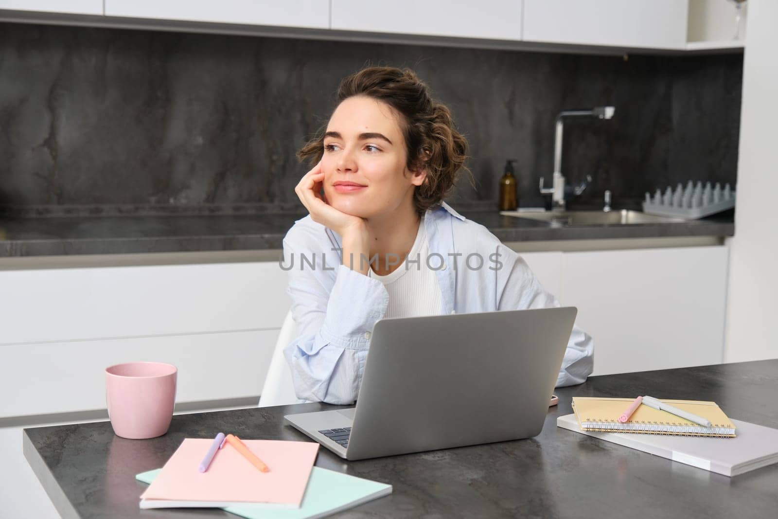 Image of young productive woman works from home, does distance learning, learns online course on her laptop in kitchen, writes down information, watches webinar, attends work meeting remotely.