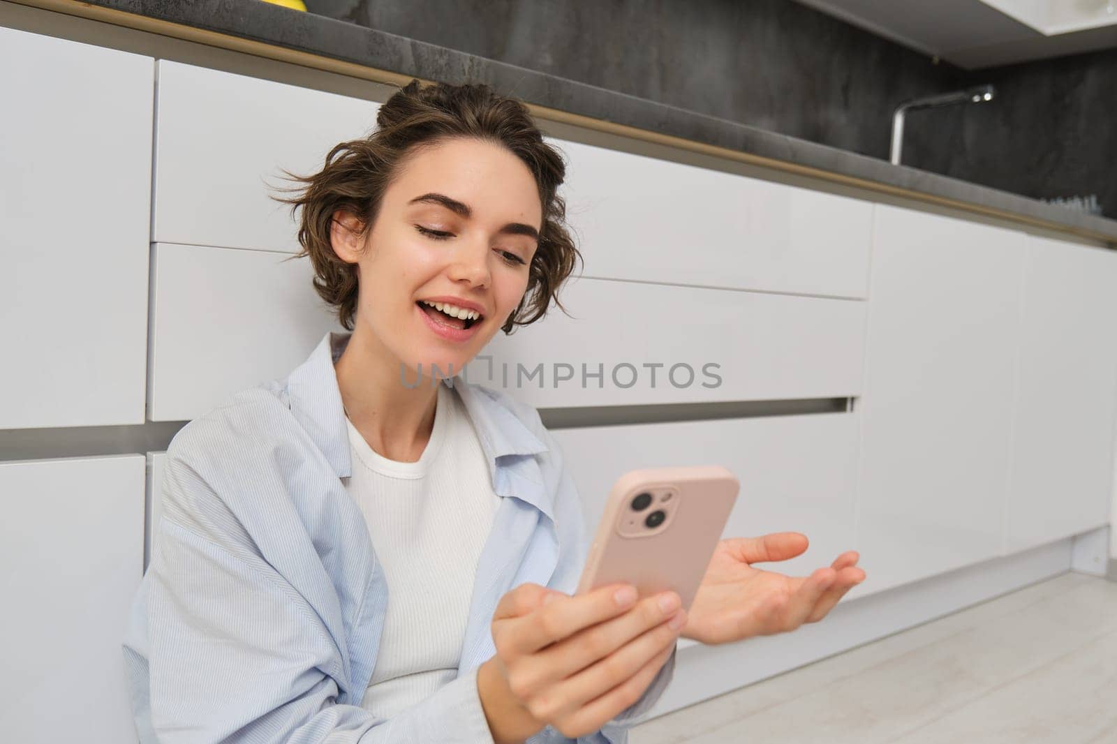 Portrait of happy young woman, shows her brand new kitchen, sits on floor, points at furniture and smiles, video chats on smartphone. Technology and lifestyle concept