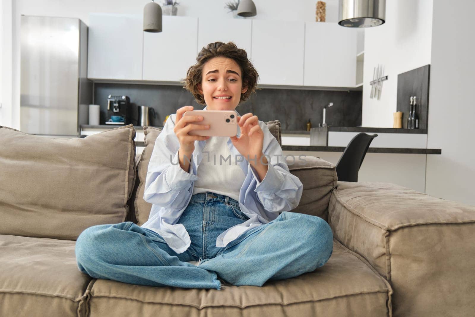 Portrait of woman playing mobile phone games, watching video with excited face, using smartphone for leisure, sits on sofa at home. Lifestyle and people concept