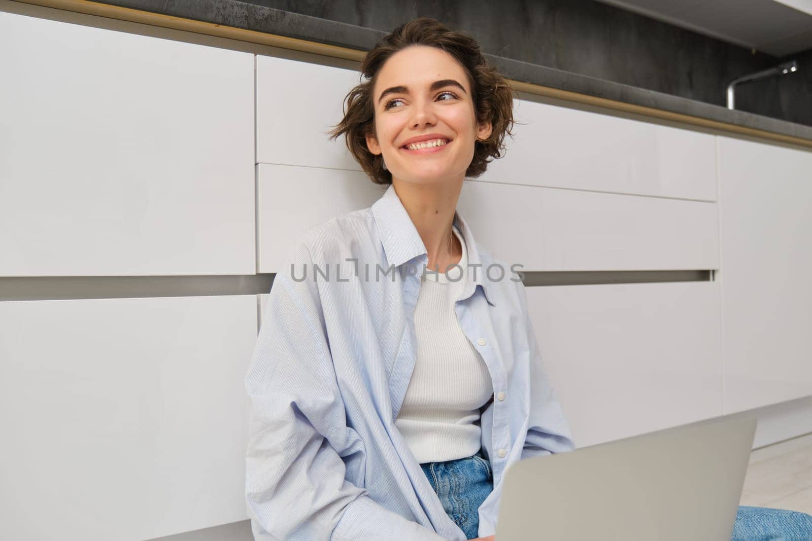 Freelance and lifestyle concept. Young woman sits on floor with laptop, works from home, sets up her remote workspace in kitchen, does homework by Benzoix