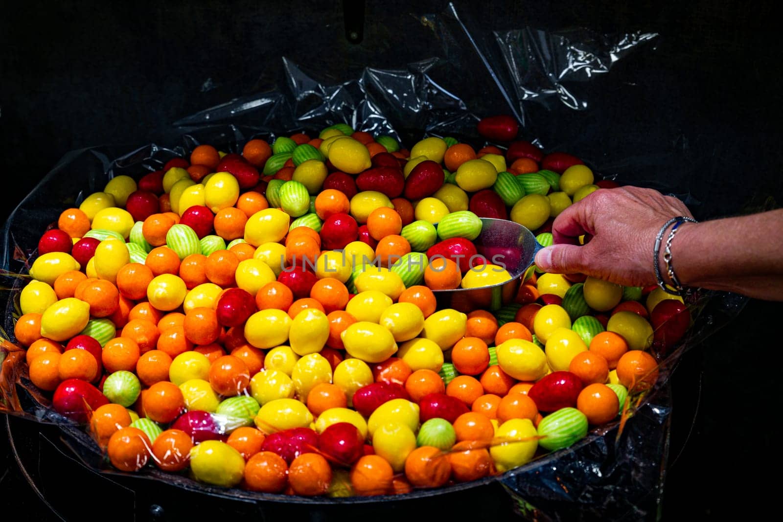 a woman's hand takes a number of candies from a large bowl with a shovel ina candy shop with an assortment of candies in orange red yellow.