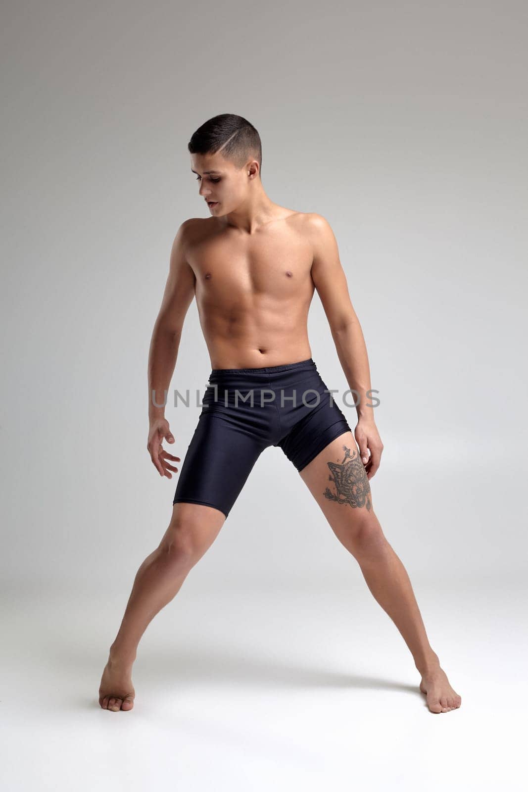 Full length portrait of a handsome muscular man ballet dancer, dressed in a black shorts. He is making a dance element against a gray background in studio. Bare legs and torso. Ballet and contemporary choreography concept. Art photo.