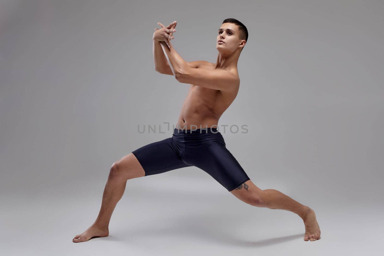 Full length portrait of an attractive handsome man ballet dancer, dressed in a black shorts. He is making a dance element with crossed hands, against a gray background in studio. Bare legs and torso. Ballet and contemporary choreography concept. Art photo.