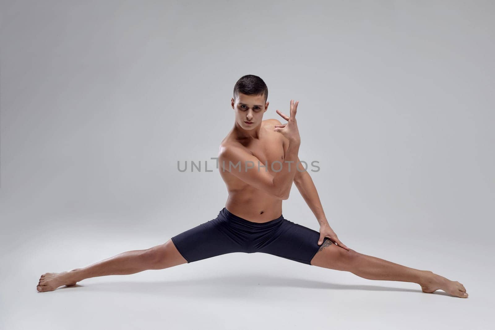 Full length portrait of a handsome muscular man ballet dancer, dressed in a black shorts. He is doing a twine against a gray background in studio. Bare legs and torso. Ballet and contemporary choreography concept. Art photo.