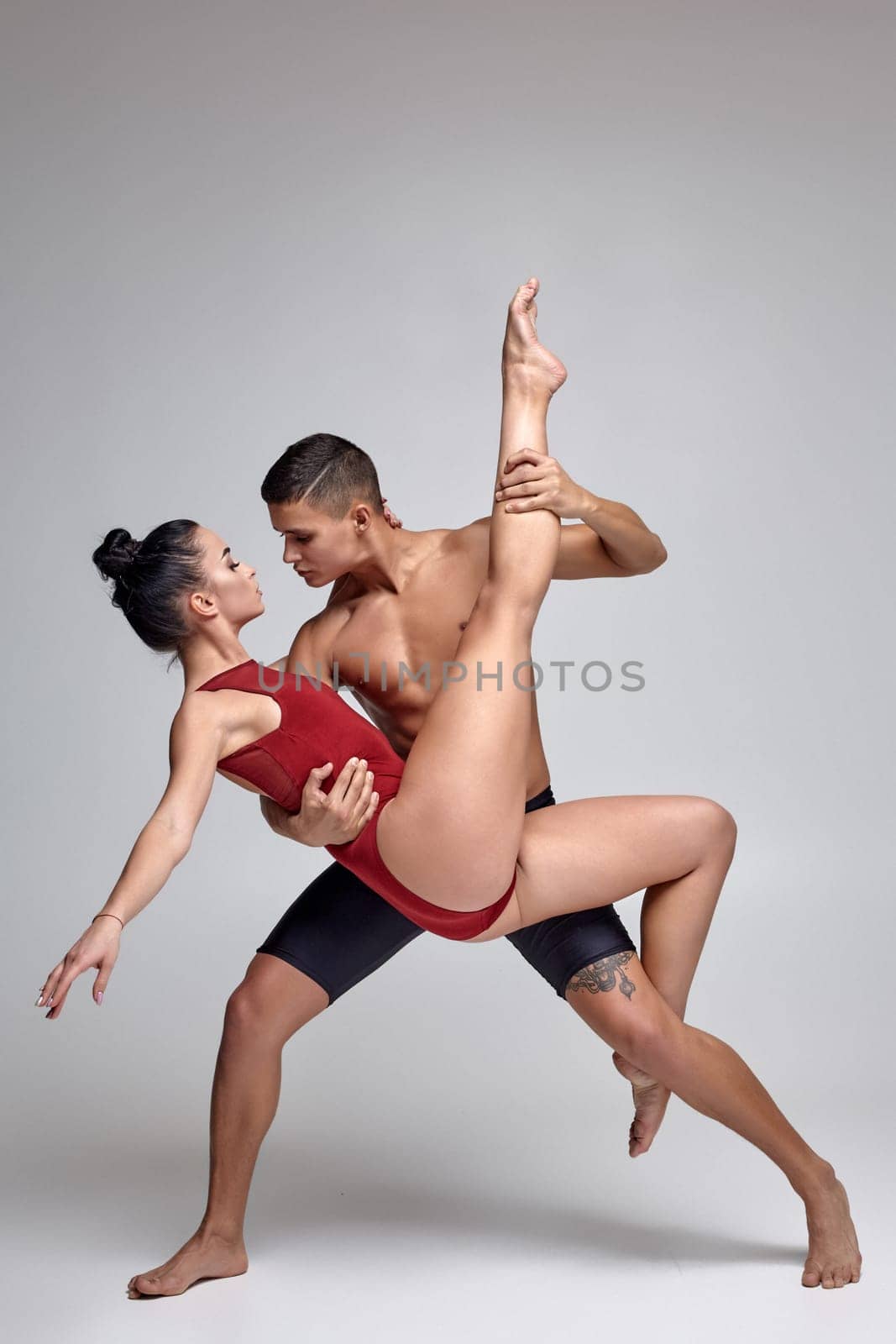 Pair of a talanted ballet dancers are posing against a gray studio background. Handsome man in black shorts and charming woman in a red swimwear are dancing together. Ballet and contemporary choreography concept. Art photo.