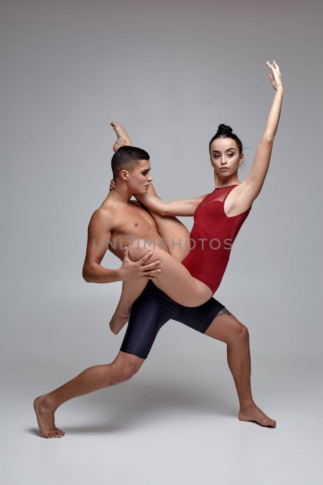 Couple of a young athletic ballet dancers are posing over a gray studio background. Handsome man in black shorts and charming woman in a red swimwear are dancing together. Ballet and contemporary choreography concept. Art photo.