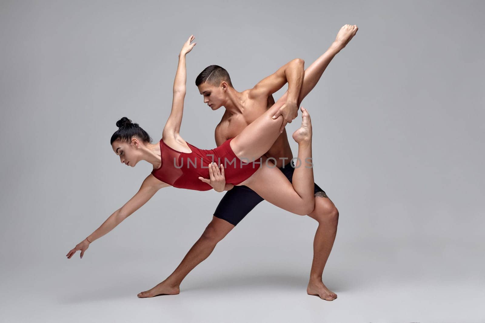 The couple of a strong ballet dancers are posing over a gray studio background. Man in black shorts and girl in a red beautiful swimwear are dancing together. Ballet and contemporary choreography concept. Art photo.