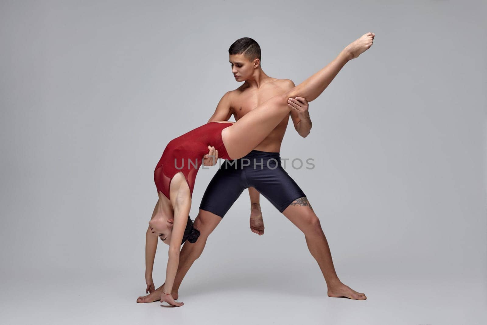 The couple of a wonderful ballet dancers are posing over a gray studio background. Male in black shorts and female in a red swimwear are dancing together. Ballet and contemporary choreography concept. Art photo.