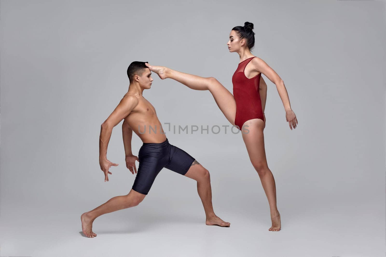 Two young modern ballet dancers are posing against a gray studio background. Good-looking man in black shorts and charming woman in a red swimwear are dancing together. Ballet and contemporary choreography concept. Art photo.