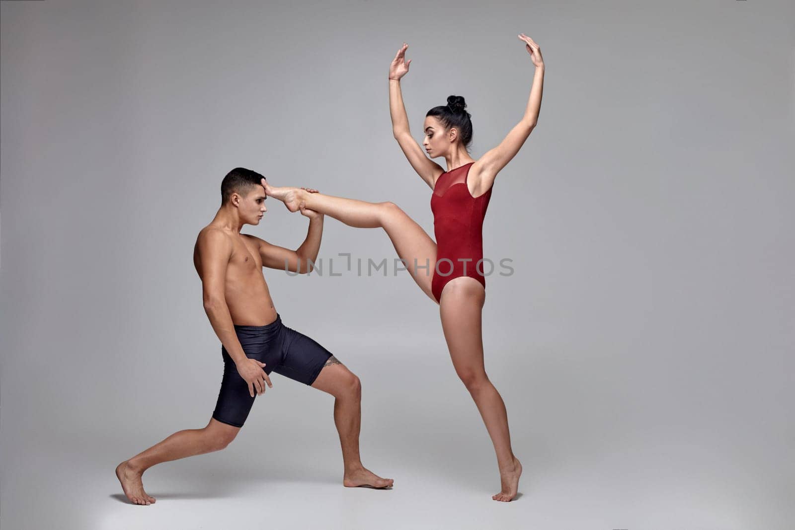 Couple of a wonderful ballet dancers are posing against a gray studio background. Handsome guy in black shorts and beautiful woman in a red swimwear are dancing together. Ballet and contemporary choreography concept. Art photo.
