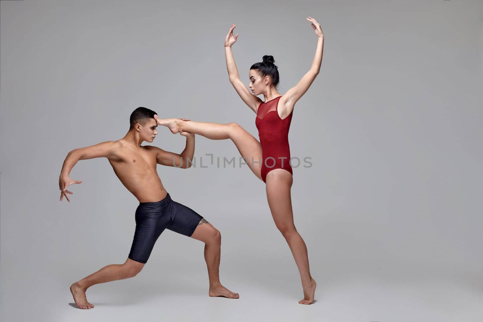 Pair of a graceful ballet dancers are posing over a gray studio background. Man in black shorts and woman in a red swimwear are dancing together. Ballet and contemporary choreography concept. Art photo.
