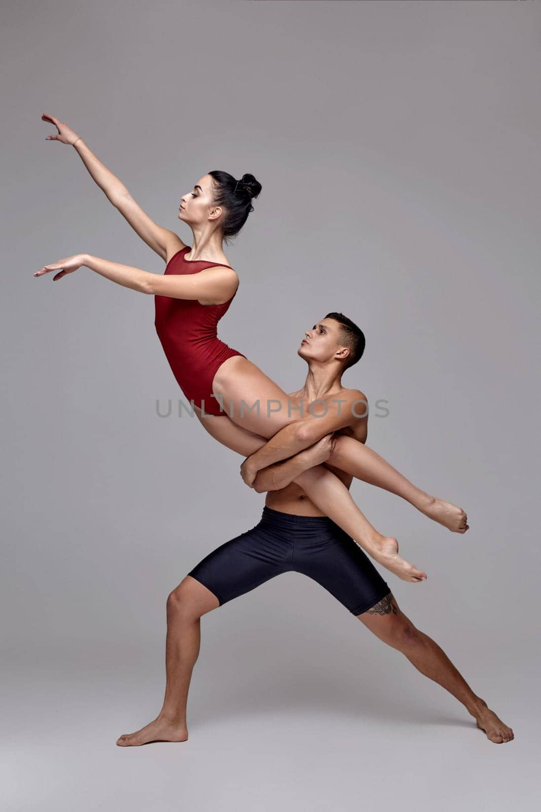 Two modern ballet dancers are posing against a gray studio background. Strong guy in black shorts and tiny girl in a red swimwear are dancing together. Ballet and contemporary choreography concept. Art photo.