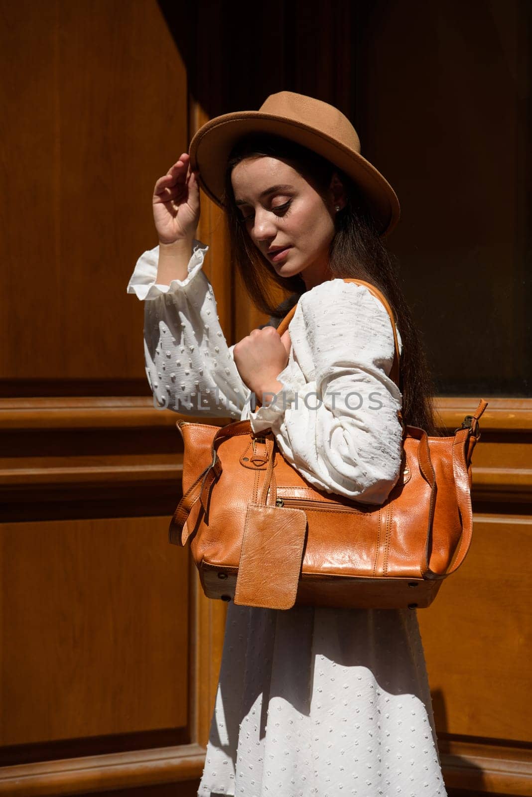 brunette with long hair wearing white dress and beige hat posing with luxury leather bag. wooden background. Vacation, tourism and travel