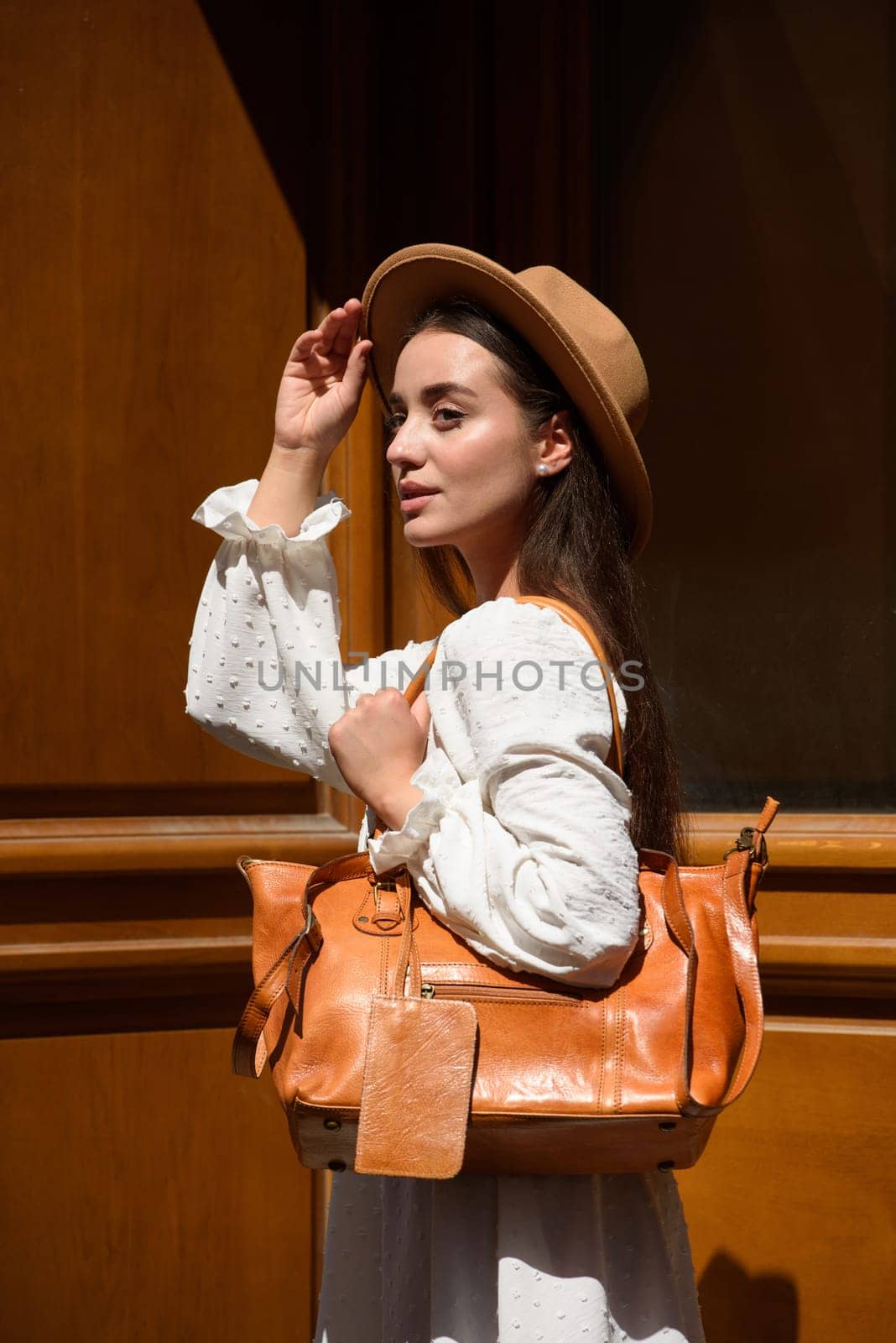 brunette with long hair wearing white dress and beige hat posing with luxury leather bag. wooden background. Vacation, tourism and travel