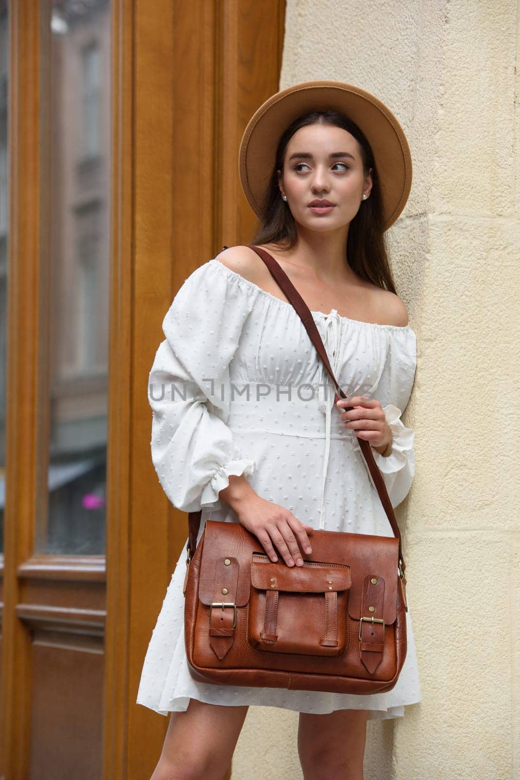 brunette with long hair wearing white dress and beige hat posing with luxury leather bag. wooden background. Vacation, tourism and travel