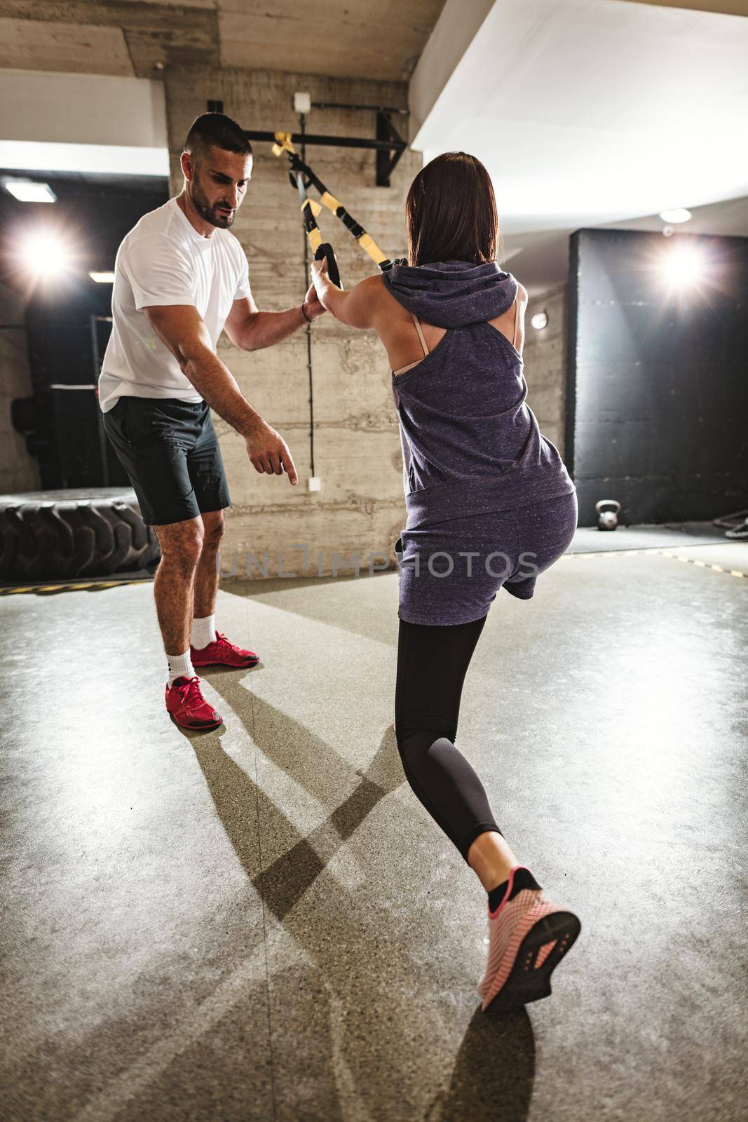 Rear view of young handsome woman having workout with a suspension straps at the gym supported of her personal male trainer. 