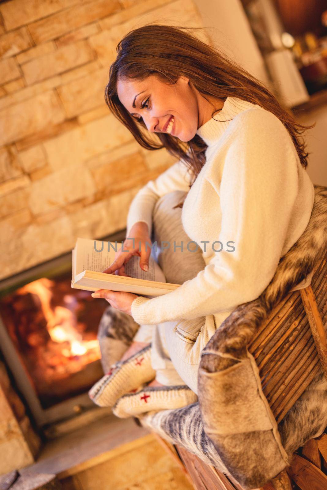 Beautiful young smiling woman reading book and enjoying by the fireplace.