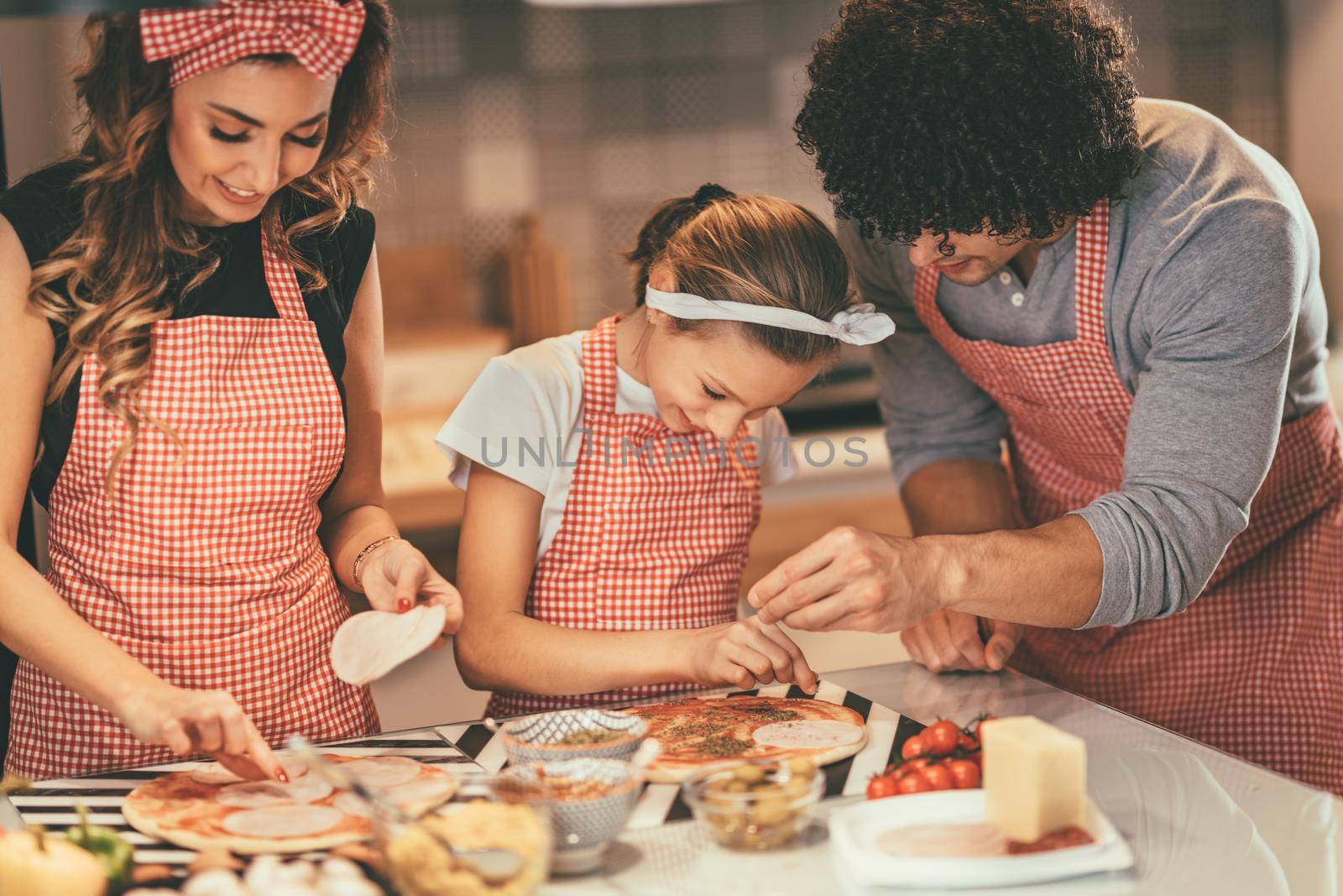 Happy parents and their daughter are preparing meal together in the kitchen. Little girl and her father are putting ketchup and oregano on the pizza.