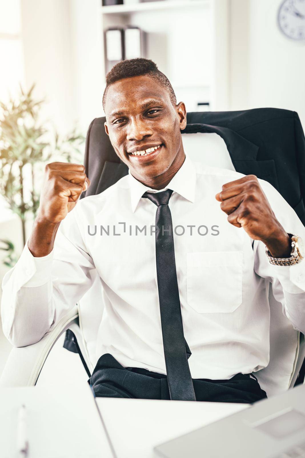 Successful African businessman celebrating success with fists in the office and looking at camera.