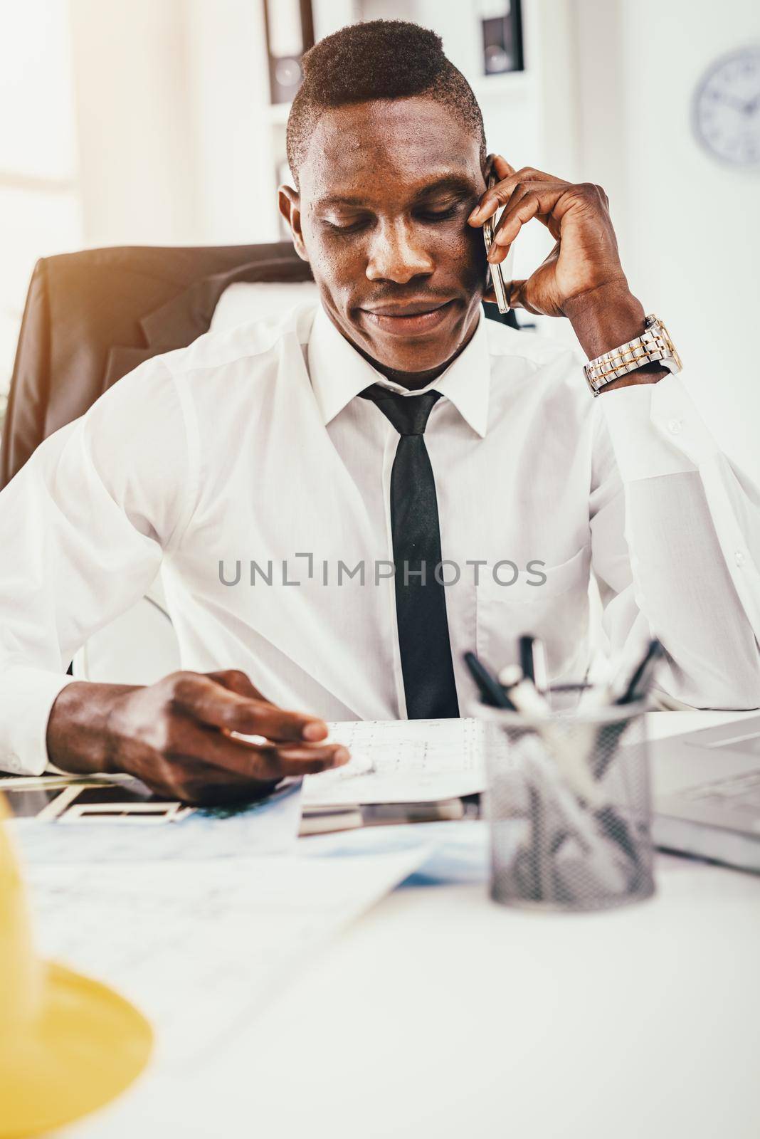 Pensive African architect talking on smartphone, analyzing blueprint and planning what to do next.