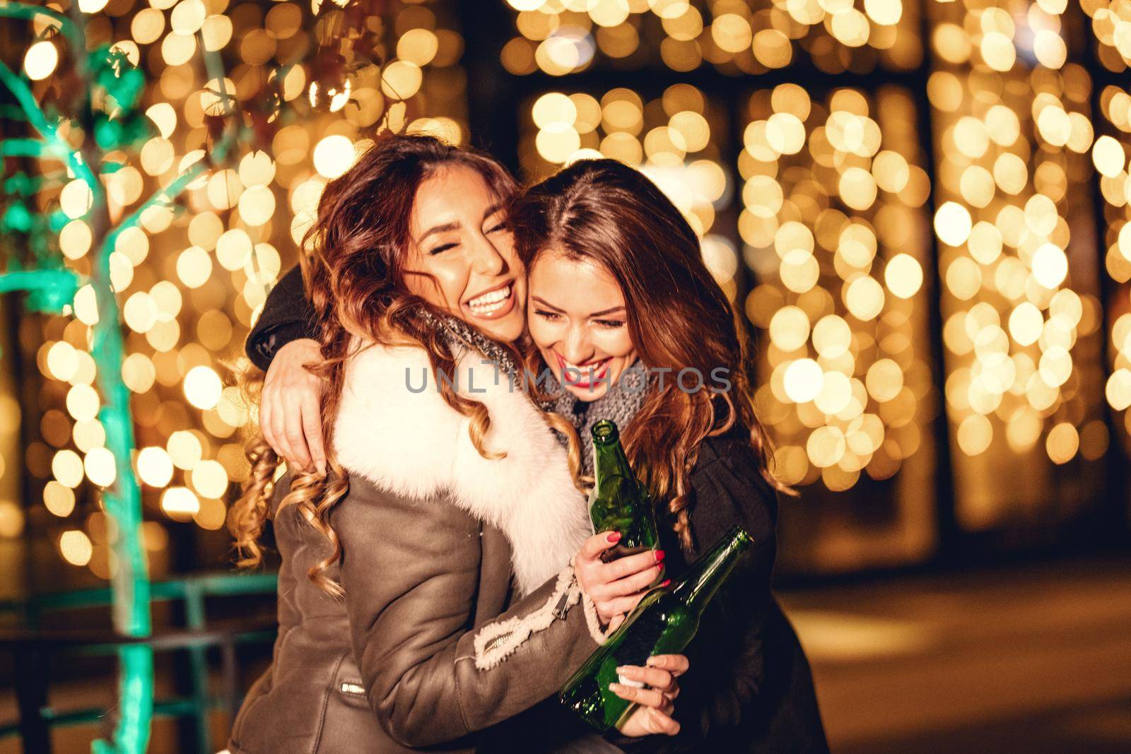 Two female friends enjoy the night out hugging and laughing with bottles of beer in their hands.