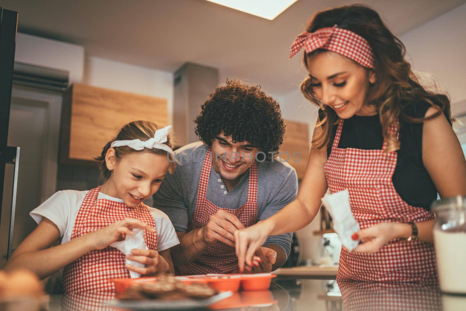 Happy parents and their daughter are preparing cookies together in the kitchen. Little girl helps to her parents to put crumbs of chocolate on the cookies.