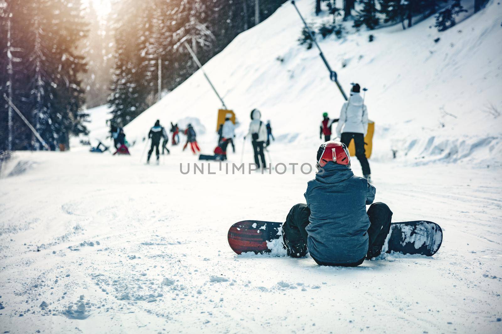 Young man with snowboard, is sitting on snow and enjoying a sunny winter day in mountains.